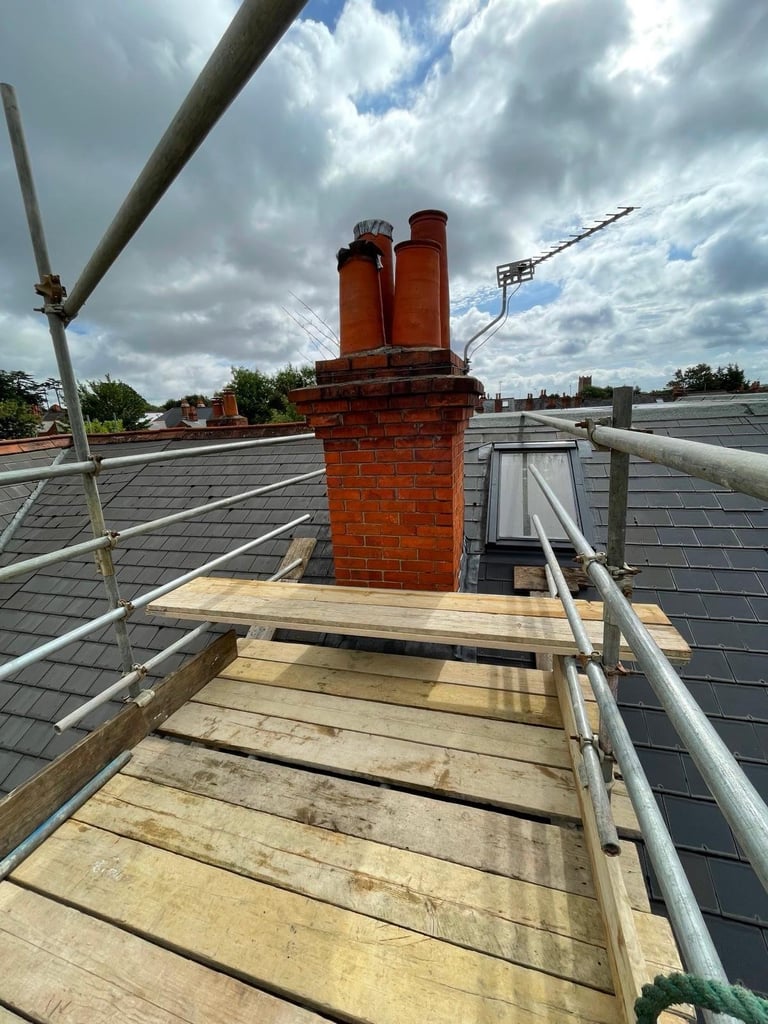 Red brick chimney on residential rooftop with wooden walkway and metal railings, partly cloudy sky