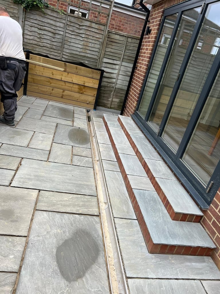 Outdoor patio with light gray stone paving, brick border edges, modern glass doors, wooden fence and planter boxes in background