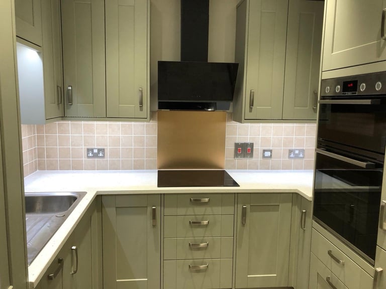 Modern sage green kitchen with white countertops, black cooktop range hood, and stainless steel oven on the right side